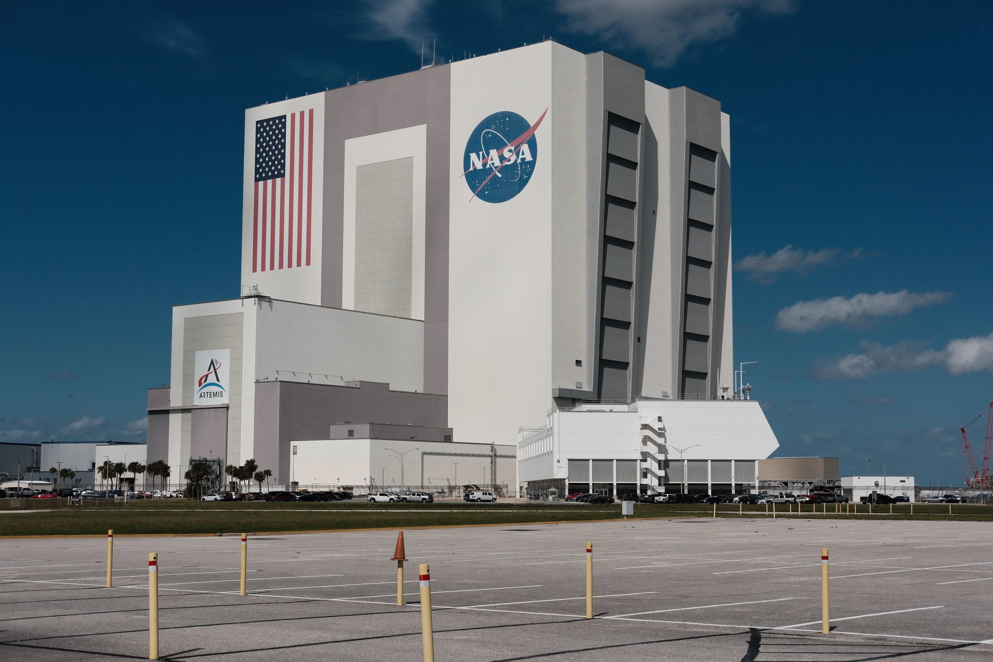 The Vehicle Assembly Building at NASA's Kennedy Space Center, its NASA insignia and American flag facing an empty visitor parking lot under a clear Florida sky